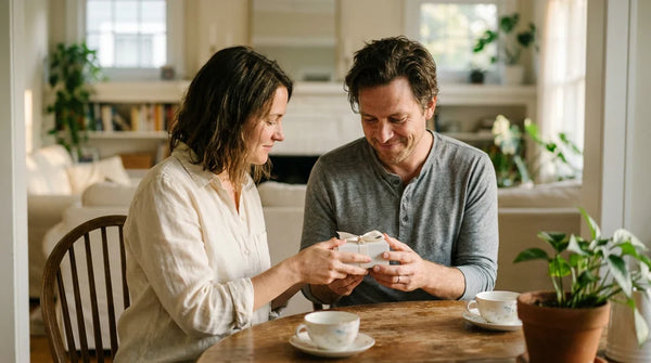 A husband and wife seated together at a wooden dining table, the wife hands him a small white jewellery gift box, soft golden hour light in their home