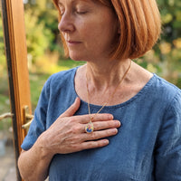 Thumbnail for Woman in a blue linen shirt with her hand on her heart, wearing the gold secret drop photo necklace | SKU: TPH-DRPN-GOLD